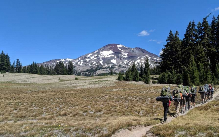 A group of outward bound students carrying backpacks hike along a trail through an open field. There is mountain spattered with snow in the background. 